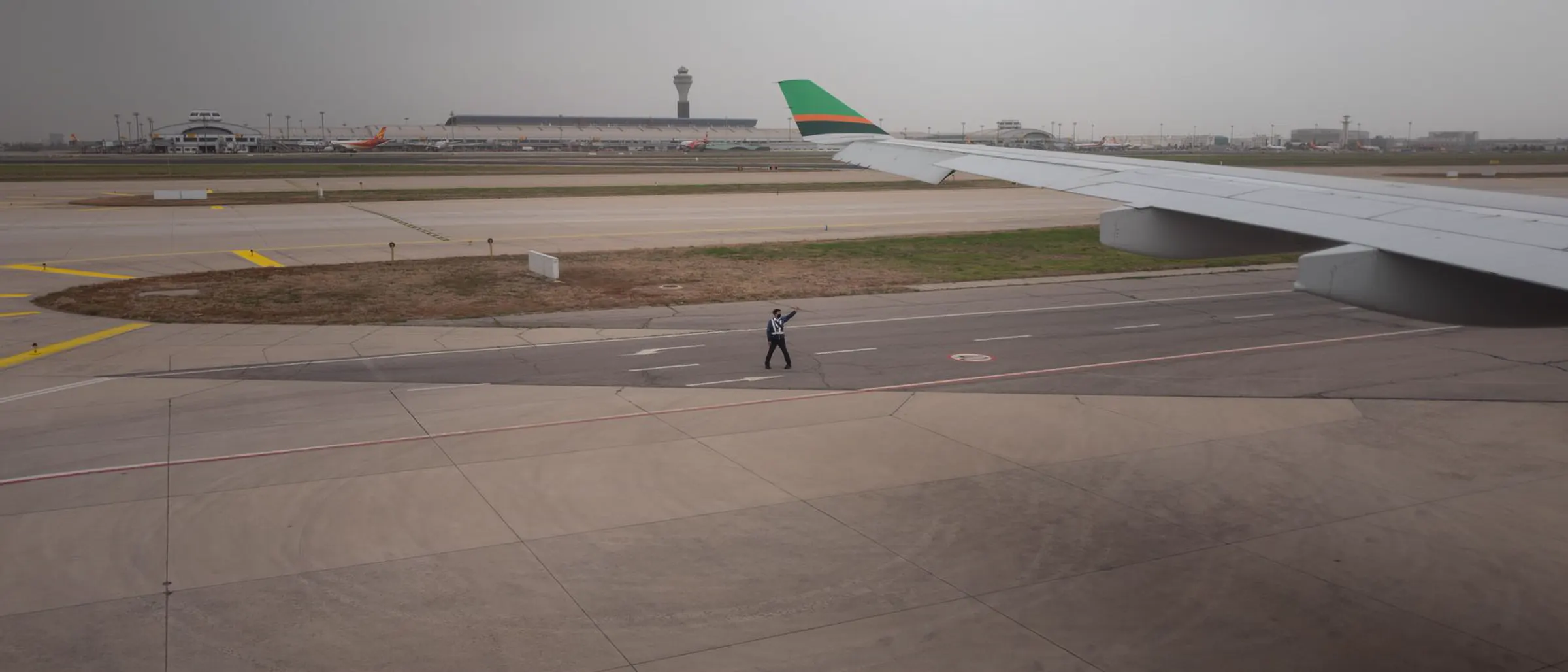 An airport apron seen past an aircraft wing, with the control tower and ground staff at work beyond it.