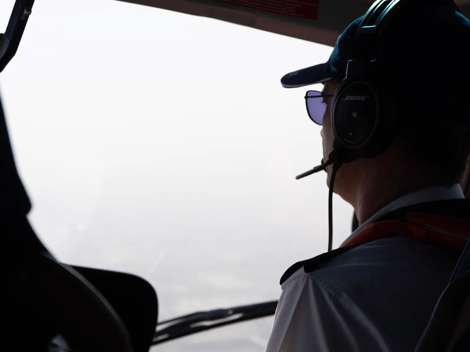 A pilot in a headset and cap seen from behind in the cockpit, turned towards a bright window.
